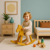 Kind lacht terwijl het speelt op een geel houten hobbelpaard in een Japandi kinderkamer met rustig interieur en Montessori speelgoed