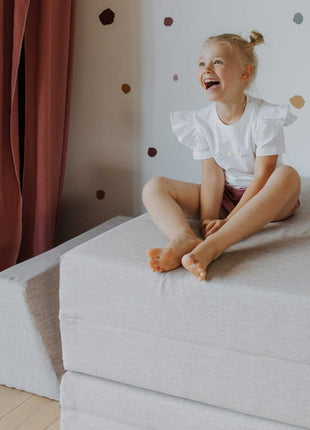 Kind zit op gestapelde matrasdelen van de beige Monboxy Montessori speelbank – ideaal voor rustmomenten en vrij spel.