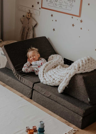 Lachend kind ligt onder een deken op de Monboxy Montessori speelbank in een comfortabele opstelling – warm en huiselijk.