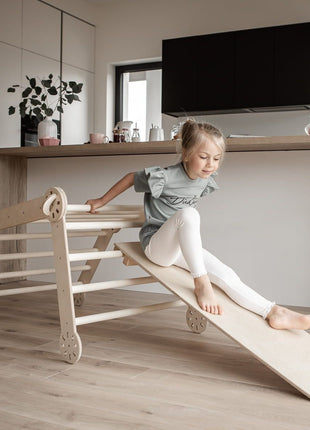 Meisje glijdt van de helling van de Montessori klimdriehoek – stimuleert zelfstandige beweging en actieve motorische ontwikkeling in huiselijke omgeving.