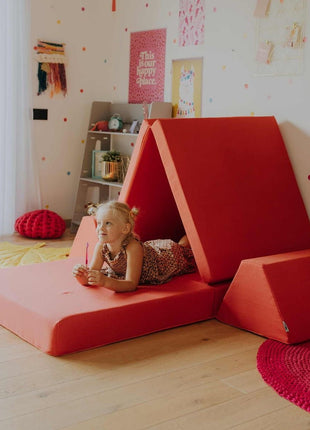 Kind ligt ontspannen in een tentvormige constructie van de koraalkleurige Monboxy Montessori speelbank – bevordert creatief spel.