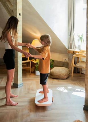 Kind balanceert op rechthoekig Leg&Go balansbord terwijl volwassene ondersteuning biedt binnenshuis