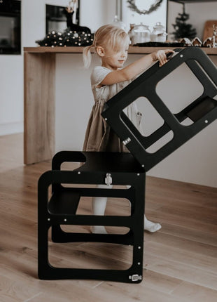 Meisje zet zwart model van Montessori 2-in-1 Keukentoren op elkaar in keukenruimte.