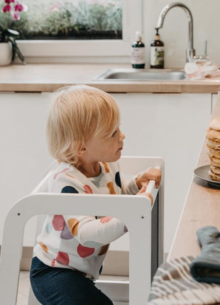 Montessori Keukenhulp met Schoolbord in wit waarop kind in keuken naar stapel pannenkoeken kijkt.