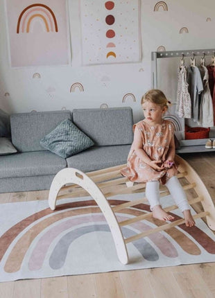 Kind zit bovenop de Montessori Klimboog op een vloerkleed met regenboogmotief in een kinderkamer.