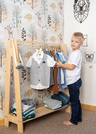 Jongen hangt kleding op Montessori Peuterkledingrek in blank hout met blauwe outfits.