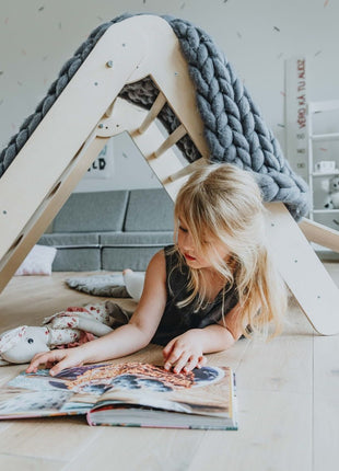 Kind ligt onder de klimdriehoek met een deken als speeltent – stimuleert rust en fantasierijk spel.