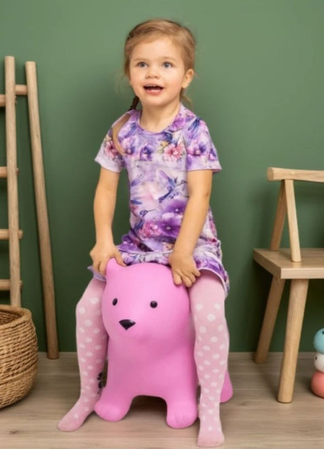 Kind zit op My First Jumpy roze springbeer bij groene muur en houten meubel.