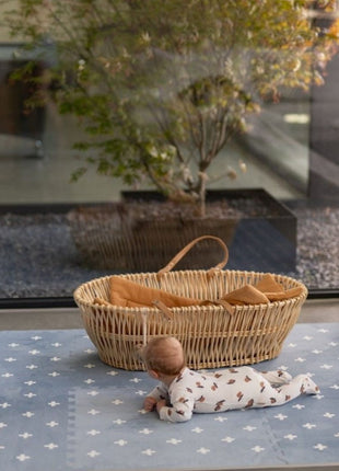 Baby ligt op de OLYA KIDS Cross Azure speelmat naast een rotan wieg bij raam.