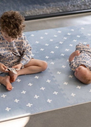 Twee kinderen op de OLYA KIDS Cross Azure speelmat in een lichte kamer met betonnen vloer.