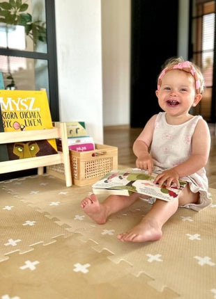 Baby zit op de OLYA KIDS Cross Sand speelmat en leest een boekje in een kinderkamer.