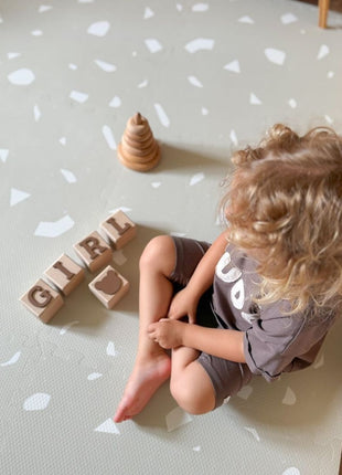 Kleuter zit op de OLYA KIDS Terrazzo Dove speelmat met houten blokken en stapeltoren.