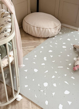 Baby kruipt op de OLYA KIDS Terrazzo Sage speelmat naast wieg en pluche kussen.