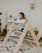 Pikler-driehoek – Pastel van leg&go met kind dat binnenshuis op de houten spijlen klimt.