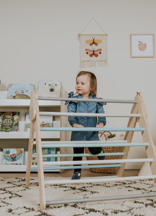 Pikler-driehoek – Pastel van leg&go met kind dat binnenshuis tussen de houten spijlen staat.