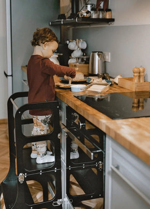 Kind smeert brood aan het keukenblad staand in de STEP'n'SIT® 2-in-1 keukentoren zwart